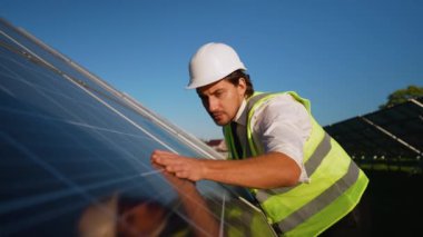 Güvenlik yeleği ve miğferi olan bir mühendis, yenilenebilir enerji alanının kalitesini ve işlevselliğini sağlamak için güneş panelinin yüzeyini dikkatle inceler.