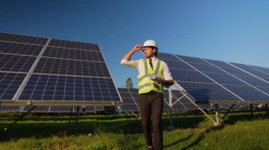 Sert şapka ve güvenlik yeleği giyen bir mühendis, parlak mavi gökyüzü altında büyük bir güneş paneli kurulumunu gözlemlerken proje planlarını gözden geçirir.