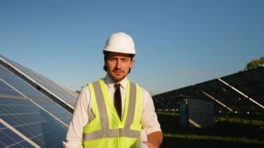 Sert şapka ve güvenlik yeleği olan bir yatırımcı güneş panellerinin yanında yürüyor, parlak ve güneşli bir gökyüzü altında yenilenebilir enerji tesisatını inceliyor.