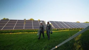 Miğfer ve üniformalı iki işçi, yenilenebilir enerji istasyonunun bakımını ve verimliliğini sağlamak için bir güneş paneli alanından araçlar ve bir merdiven taşıyarak yürüyorlar.