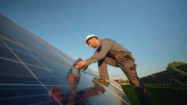 Tulum ve miğfer giyen profesyonel bir işçi güneş panelini emniyete almak için matkap kullanır. Kurulumun optimal enerji performansı için güvenli ve verimli olduğundan emin olur.