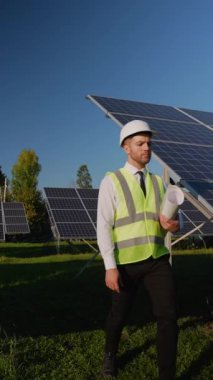 Güvenlik yeleği ve miğferinde profesyonel mühendis berrak mavi gökyüzünün altındaki yenilenebilir enerji alanındaki güneş panellerini inceliyor.