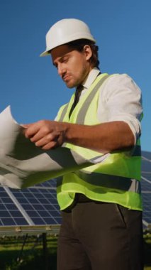 Sert şapka ve güvenlik yeleği giyen bir mühendis, güneş panellerinin yanında dururken projenin özellikleriyle açık bir gökyüzü altında aynı hizayı sağlayarak projeleri gözden geçirir.