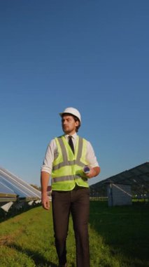 Kask ve güvenlik yeleği giyen bir mühendis, güneş enerjisi alanındaki operasyonları denetlerken proje planları tutuyor.