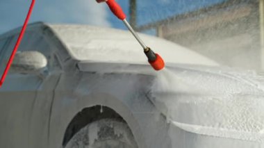 Açık hava oto yıkama servisinde arabasını köpükle kaplamak için kırmızı basınçlı çamaşır makinesi kullanan bir adam güneşli bir günde temizlik yapar.