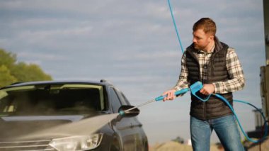 Arabalarını temizlemek için yüksek basınçlı su hortumunu kullanan bir adam, açık hava ve parlak güneş ışığı altında, açık hava oto yıkama servisinde lekesiz bir son garantiliyor.