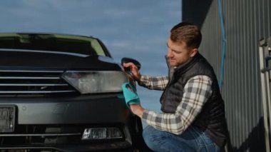 Turkuaz mikrofiber bez kullanarak arabasının farlarını temizlerken gülümseyen bir adam güneşli bir günde açık hava oto yıkama servisinde cilalı bir görünüm sağlıyor.