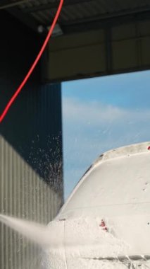 Sabun köpüğüyle kaplı bir arabayı temizlemek için basınçlı çamaşır makinesi kullanan, günlük kıyafetler giyen ve güneşli bir günde açık hava araba yıkama yerinde modern bir aracın yanında duran bir adam.