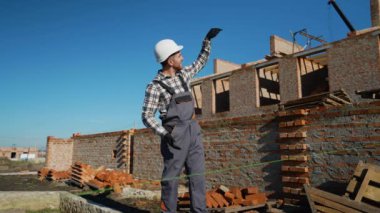 Bir inşaat denetçisi, parlak mavi gökyüzünün altındaki tuğla şantiyede inşaat sürecini denetlerken güvenlik vitesi jestleri yapıyor.