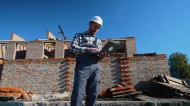 Güvenlik teçhizatı kullanan bir inşaat ustası, inşaat verilerini bir tuğla inşaat sahasındaki dizüstü bilgisayara yerleştiriyor.