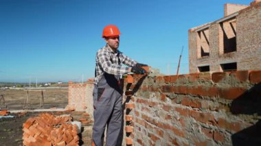 Güvenlik teçhizatı içinde profesyonel bir inşaat işçisi bir kulübe inşa ederken dikkatli bir şekilde harçla tuğla döşeyerek beceri ve hassasiyeti ön plana çıkarıyor.
