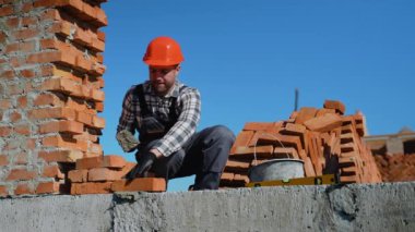 Güvenlik donanımlı bir inşaat işçisi dikkatli bir şekilde tuğla döşeyip, parlak mavi gökyüzünün altında aktif bir inşaat alanı inşa ederek, işçiliğini sergiliyor.
