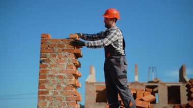 Güvenlik teçhizatı takan yetenekli bir tuğla ustası, açık mavi gökyüzünün altında etkin bir inşaat alanında duvar inşa ederken tuğlalara harç uygular.