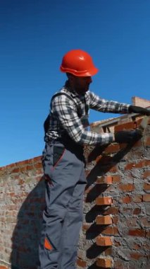 Güvenlik donanımlı bir inşaat işçisi dikkatli bir şekilde tuğla döşeyip, parlak mavi gökyüzünün altında aktif bir inşaat alanı inşa ederek, işçiliğini sergiliyor.