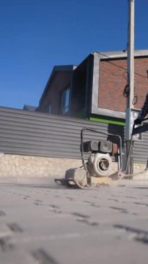 Bir işçi, yerleşim bölgesinde yeni döşenmiş kaldırım taşları üzerinde plaka sıkıştırıcı kullanıyor, açık gökyüzünün altında koruyucu giysiler giyiyor.