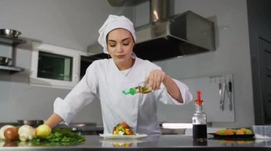 Beyaz üniformalı profesyonel bir kadın şef taze yapılmış sebze salatasının üzerine dikkatlice yağ dökerek mutfak sanatını ve sağlıklı mutfağı ön plana çıkarıyor..
