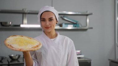 Beyaz üniformalı neşeli bir şef, profesyonel bir mutfak ortamında üzerinde dilimlenmiş peynir olan pişmemiş bir pizzayı elinde tutuyor..