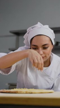 Beyaz üniformalı bir şef, profesyonel bir mutfak ortamında pizza hamuruna dilimlenmiş peynir ekliyor. Pişirmeye hazırlanıyor..