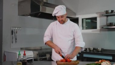 Beyaz üniformalı profesyonel bir aşçı ahşap bir tahtada taze domates dilimliyor, modern bir mutfakta yemek için malzemeler hazırlıyor..