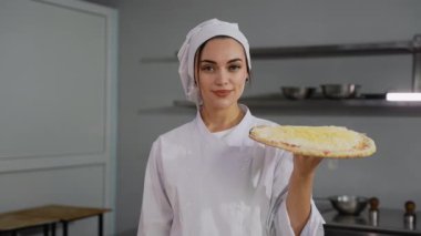 Beyaz üniformalı profesyonel bir şef, üzerinde peynir olan pişmemiş bir pizzayı, temiz bir mutfak ortamında kendine güveniyor..