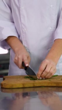 Şefin elleri beyaz bir üniformayla profesyonel bir mutfakta ahşap bir kesme tahtasındaki taze yeşil otları keserken..