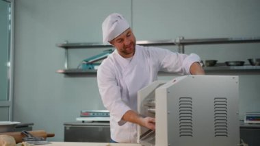 Beyaz üniformalı profesyonel bir aşçı gülümseyen bir makina kullanıyor, ticari bir mutfakta çalışıyor..