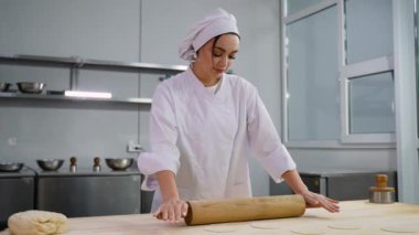 Beyaz üniformalı bir kadın aşçı profesyonel bir mutfak ortamında hamur düzleştirmek için ahşap bir oklava kullanıyor..