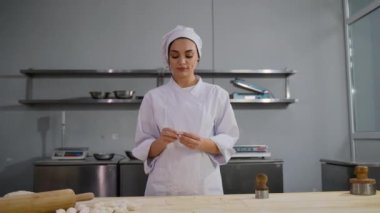Beyaz üniformalı bir şef ahşap bir yüzeye elle hamur ambalajlarını katlıyor ve arka planda mutfak ekipmanları var..