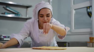 Beyaz üniformalı bir şefin, profesyonel bir mutfak ortamında pizzaya peynir serptiği yakın plan bir fotoğraf..
