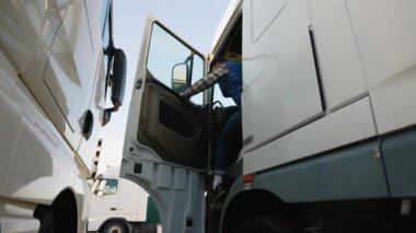 Mavi yelek ve ekose gömlekli bir kamyon şoförü park halindeki bir kamyonun kabininden çıkıp, park yerinde mola veriyor.