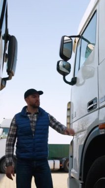 Mavi yelek ve ekose gömlekli bir kamyon şoförü park halindeki bir kamyonun kabinine giriyor, açık bir otoparkta kalkışa hazırlanıyor.