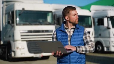 Mavi yelekli ve ekose gömlekli bir depo işçisi kamyon yükleme işini denetlemek ve yönetmek için dizüstü bilgisayar kullanıyor.