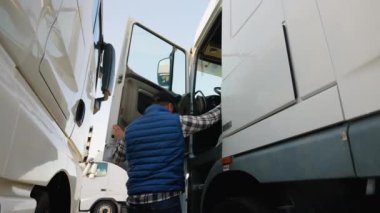 Mavi yelek ve ekose gömlekli bir kamyon şoförü park halindeki bir kamyonun kabinine giriyor, açık bir otoparkta kalkışa hazırlanıyor.