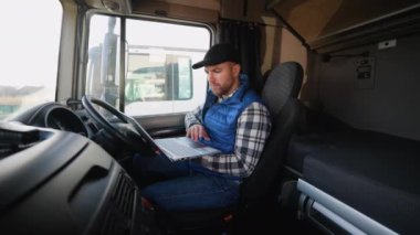 Mavi yelek ve ekose gömlekli bir kamyon şoförü, park halindeki kamyonların yakınındaki gezi belgelerini incelemek için dizüstü bilgisayar kullanıyor.
