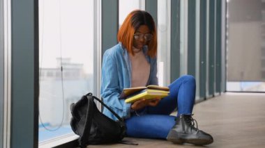 Gözlüklü ve kot ceketli genç Afrikalı bir kadın büyük bir pencerenin yanında oturmuş sırt çantasıyla kitap okumaya odaklanmış..