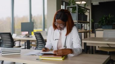 Genç Afrikalı Amerikalı kadın gözlüklü, beyaz önlüklü ve steteskoplu, modern bir üniversite kütüphanesinde kitaplarla çalışırken notlar yazıyor..