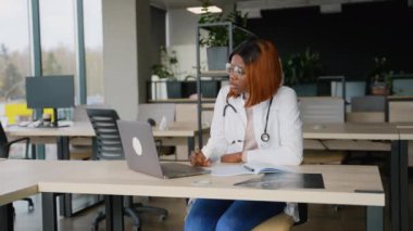 Beyaz laboratuvar önlüğü ve steteskop giyen genç Afrikalı bir tıp öğrencisi modern bir üniversite kütüphanesinde dizüstü bilgisayarla internette tıp okuyor..