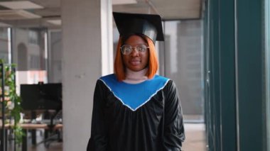 Siyah kep ve mavi kuşaklı bir cübbe giyen Afro-Amerikan mezunu. Eğitimini tamamladıktan sonra gururlu ve kendinden emin bir ifadeyle içeride duruyor..