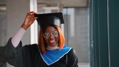 Mavi kuşaklı siyah bir cübbe ve cübbe giyerek mezun olan mutlu Afrikalı Amerikalı, kapalı alanda gururla gülümserken mezuniyet şapkalarını düzeltiyor..