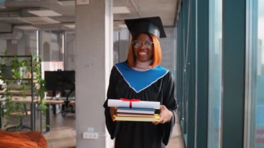 Siyah şapkalı ve cüppeli gülümseyen bir mezun gururla elinde kırmızı kurdele ve bir yığın kitapla akademik başarısını kutlayan bir diploma tutuyor..