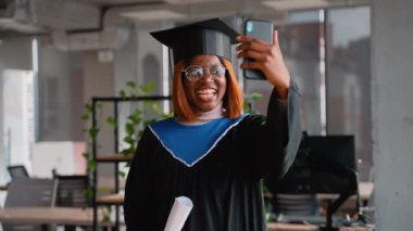 Şapka ve cüppe giyen neşeli bir mezun mezuniyet töreninden sonra ailelere kutlama videosu çeker, heyecan ve başarıyı paylaşır..