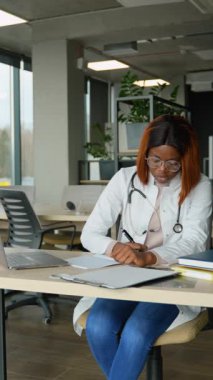 Beyaz önlüklü, steteskoplu Afro-Amerikan tıp öğrencisi, modern bir çalışma alanındaki bir dizüstü bilgisayardan öğretmeniyle iletişim kurarken notlar alıyor..