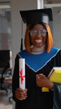 Siyah şapkalı ve cüppeli gülümseyen bir mezun gururla elinde kırmızı kurdele ve bir yığın kitapla akademik başarısını kutlayan bir diploma tutuyor..