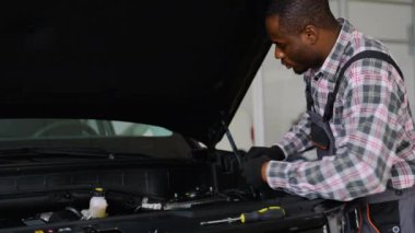 Tulumlu ve eldivenli Afrikalı bir otomobil tamircisi, bakım, tanı ve teftiş için araç gereçleri kullanarak bir atölyedeki araba motorunu tamir eder..