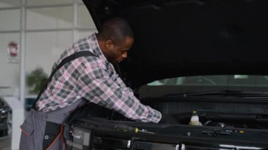 İş tulumu ve eldiven giyen profesyonel bir oto tamircisi, tamirat ve bakım üzerine çalışan bir atölyede araba motoru üzerinde çalışır..