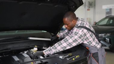 Eldiven ve tulumlu Afrikalı bir oto tamircisi modern bir atölyede çalışma lambasını kullanarak bir araba motorunu inceler, teşhis ve onarımlara odaklanır..