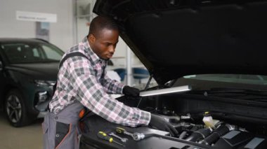 Eldiven ve tulum giyen profesyonel bir oto tamircisi atölyede bir araba motorunu kontrol eder ve onarım ve tanı konularına odaklanır..