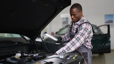 Tulum ve eldiven giyen Afrikalı bir oto tamircisi modern bir atölyede tamire başlamadan önce çalışma lambasıyla bir araba motorunu inceler..