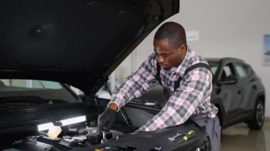 Eldivenli ve tulumlu Afrikalı bir oto tamircisi modern bir atölyede çalışma lambasının altındaki bir araba motorunu onarıyor, teşhis ve bakım için araçlar kullanıyor..