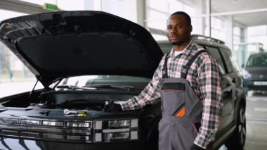 Tulumlu ve eldivenli bir Afrikalı Amerikalı oto tamircisi modern bir atölyede kaputu açık, bakım için hazır bir arabanın yanında duruyor..
